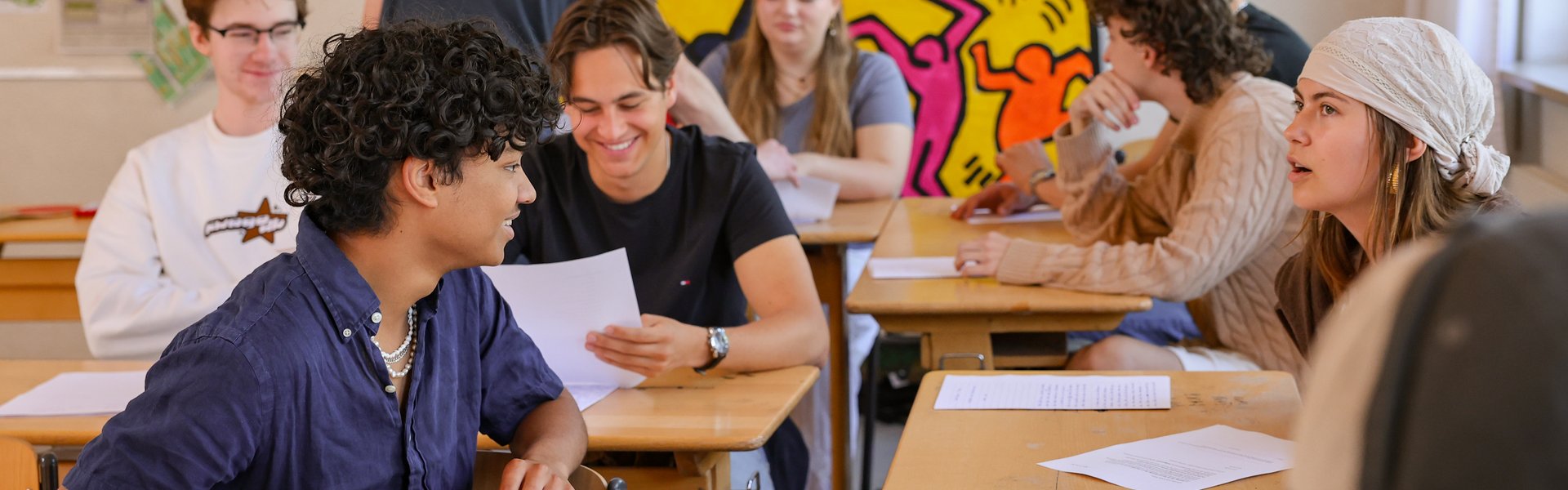 Schüler sitzen an den Tischen in einem Klassenzimmer, unterhalten sich und lächeln, während sie ihr Arbeiten anschauen. Vorne steht ein Lehrer. Helle Kunstwerke mit tanzenden Figuren zieren die Rückwand. Die Stimmung ist lebhaft und einnehmend.