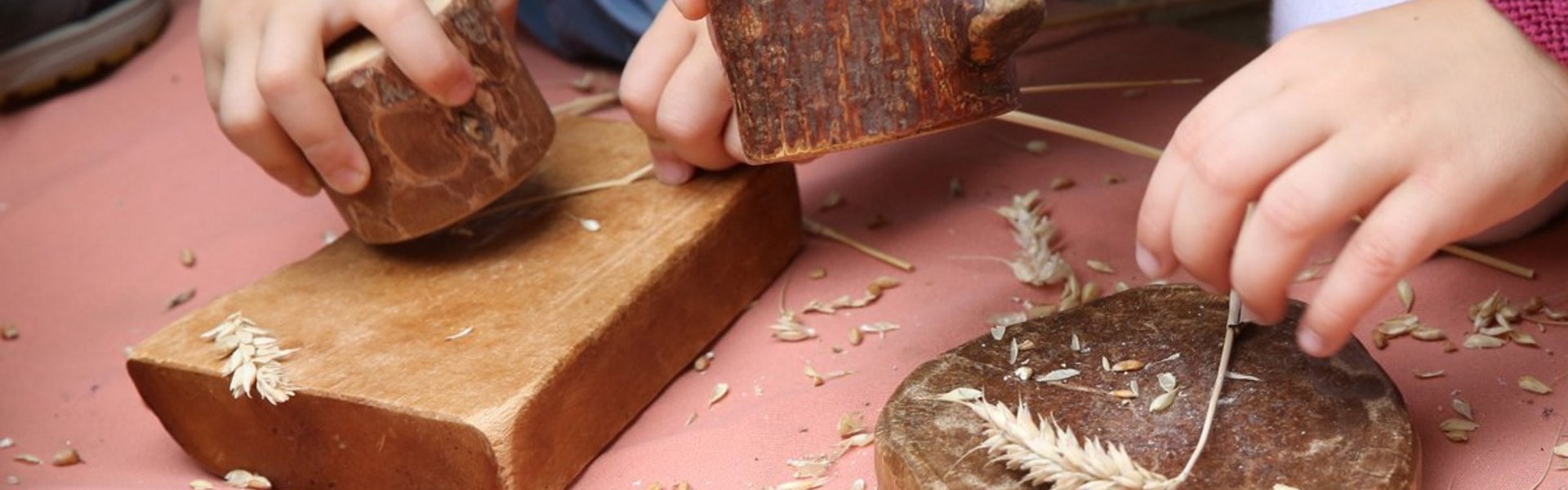 Zwei Kinder mahlen mit Holzwerkzeugen Weizenköpfe auf flachen Holzbrettern, wobei die Weizenkörner und -spelzen auf einer rosafarbenen Fläche verstreut sind. Nur ihre Hände und Unterarme sind sichtbar.